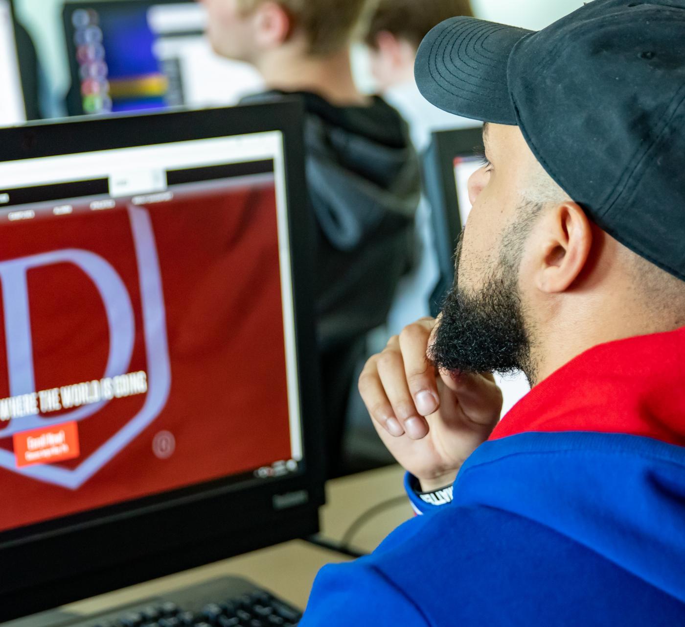 Student with a hat in class on a laptop listening to a professor